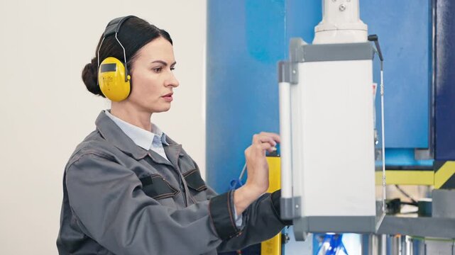 Focused Caucasian woman wearing yellow protective headphones operating industrial equipment. Carefully pressing inputs on control screen. Concentrated on adjusting machine settings inside factory.