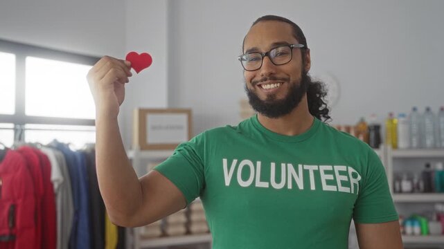 Man smiling and holding a red heart token with hand in donation center building, volunteer shirt visible; community compassion.