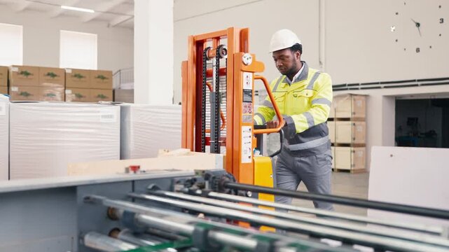 African American male factory worker wearing reflective jacket and white helmet pushing orange lifter with determination. Focused expression while moving materials in spacious warehouse with goods.