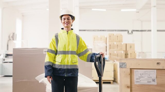 Charming Caucasian female worker wearing reflective jacket and white helmet smiling confidently. Holding handle of pallet jack while standing in bright warehouse surrounded by wooden crates.