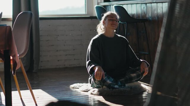 Young woman in a sweater and pajama pants meditating on the floor in the morning at a country house. Mindfulness, relaxation, slow living and cozy home wellness lifestyle concept.