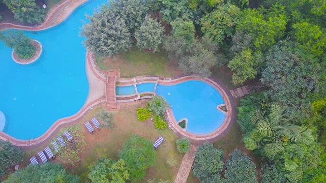 Aerial drone shot moving sideways over a huge luxurious blue pool with beach chairs on the side and a stone path running all around showing luxurious living in gurgaon, delhi, mumbai, bangalore in