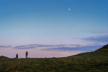 Fototapeta premium Wicklow Head dusk with two silhouetted figures, textured grass and half-moon over serene Irish Sea, autumn evening light