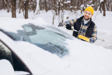 Smiling man cleaning car windshield from winter snow and ice with brush and scraper tool outdoors