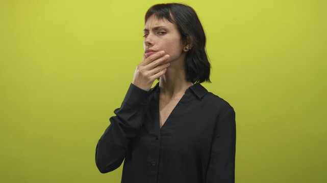 Young hispanic woman touches chin in yellow studio with slight frown and direct gaze; contemplation.