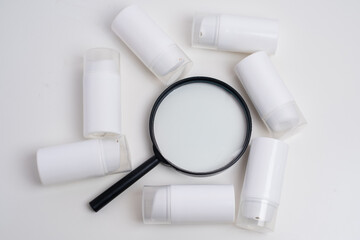 Magnifying glass focusing on white cream bottle with multiple containers in background. Cosmetic product research concept. 