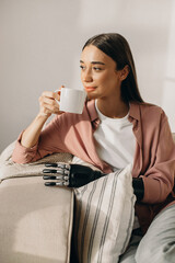 Vertical image of lovely young lady with bionic robotic hand, holding cup of coffee while resting on sofa and looking aside with pensive facial expression, trying to adapt to new artificial limb