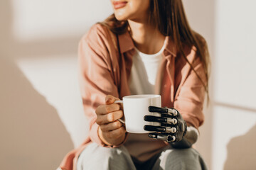 Cropped shot of unrecognizable female cyborg with bionic hand holding white mug enjoying coffee and warm sun rays coming through window into room. Robotic technologies for people with special needs