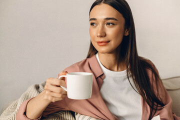 Close-up portrait of stunning gorgeous charming adorable cute lovely young woman with long hair holding white cup of coffee in hand, looking aside, admiring sunset through window in early morning
