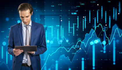 Businessman in suit using tablet device with financial data and stock exchange graphs on digital futuristic screen in blue background office.