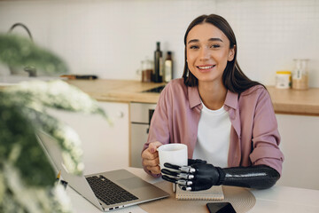 Good-looking teen girl in casual clothes holding cup of coffee by artificial bionic hand, sitting at table with laptop remote learning online. Rehabilitation and hi-tech medicine concept