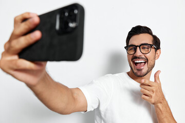 Happy smiling man in glasses takes a selfie with smartphone, showing thumbs up while standing...