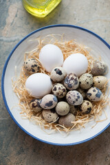 Raw quail and chicken eggs in a white and blue plate, vertical shot on a beige granite background, middle close-up, high angle view