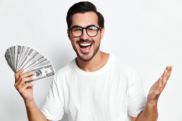 Happy smiling man holds fan of dollar bills, wearing glasses and white t-shirt, looks at camera...