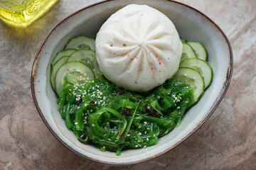 Bowl with steamed baozi, wakame seaweed salad and cucumber, horizontal shot on a brown granite background