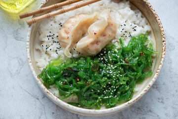 Beige bowl with rice, wakame seaweed salad and steamed dumplings, horizontal shot on a white stone background, middle closeup