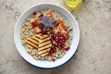 Plate with quinoa, cherry tomatoes, pomegranate seeds and grilled halloumi, horizontal shot on a beige granite background, top view