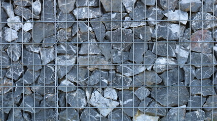 A fragment of a gabion with a metal cage filled with large gray boulders, the hard, solid texture of stone and metal, a view of a finished gabion with natural rock filling behind an iron grate © Skyliz