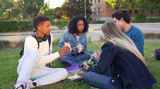 Group of diverse young friends sitting on the grass at university, enjoying their lunch break together, eating and talking happily
