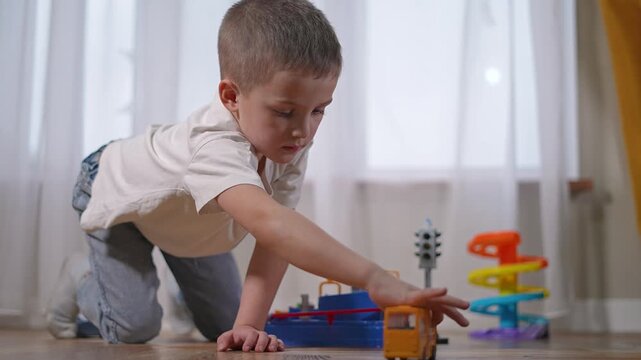 Preschooler reaches forward with bus. Child kneels near spiral slide and signal toy. Learner carefully moves bus past toy light. Boy fully focused on movement, student learning through play.