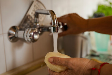 Close-up view of hands rinsing a peeled potato under running tap water in a kitchen sink. The image shows washing vegetables during food preparation in a home kitchen.