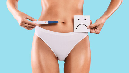 Unplanned Pregnancy. Young woman holding positive pregnancy test with two lines and sticker post-it note card with unhappy sad smiley face. Unrecognizable female in white panties. Closeup cropped view