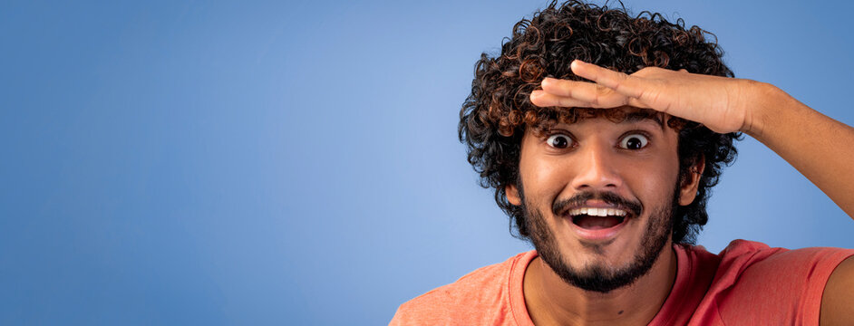 A young man stands with his hand over his forehead, looking ahead with a big smile. He has curly hair and is wearing a casual shirt. The background is blue, which adds to the cheerful mood.