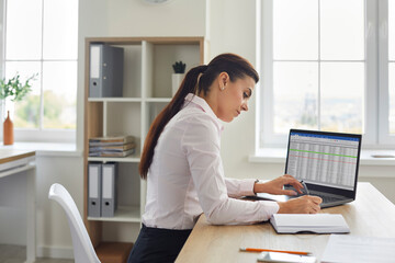 Woman with laptop at home office managing finance. She reviews accounting spreadsheets and writes in a notebook to track expenses and income. Clear concept: budgeting and personal finance.