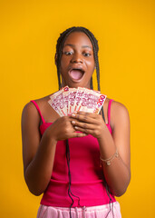 An excited young woman with braided hair and a shocked expression holds a fan of Ghanaian 200 Cedi notes. She wears a pink top against a bright yellow background, looking down at the money in awe