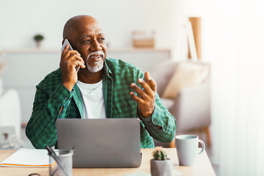 A senior man is sitting at a table, using a laptop and holding a phone to his ear. He is talking while gesturing with his other hand. The room is well-lit with soft light. - Powered by Adobe