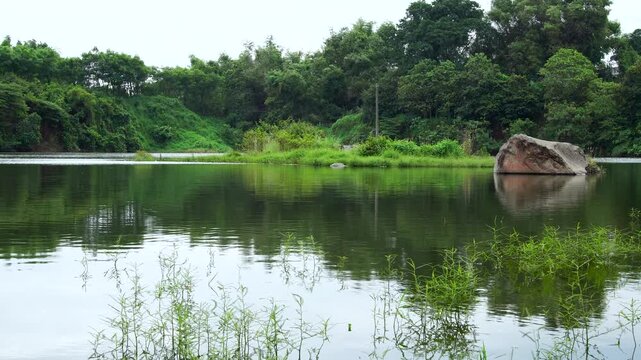 Static landscape view of Lake Ranu Manduro featuring a massive boulder rock and lush green vegetation. Serene nature scenery in Mojokerto, East Java, Indonesia.