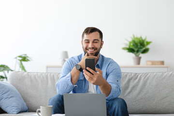 Happy middle aged european male with beard work remotely at laptop, look at phone in living room interior. Remote job, freelance, social distance, new app and gadgets. New normal, covid-19 outbreak