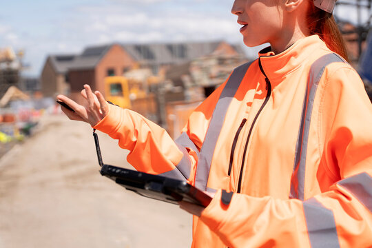 Portrait of smiling young woman builder site engineer in bright safety gear using rugged tablet at construction site
