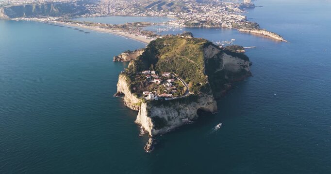 Aerial view of the rugged islet of Punta Pennata surrounded by deep blue waters, with a boat sailing nearby, Capo Miseno, Campania, Italy.