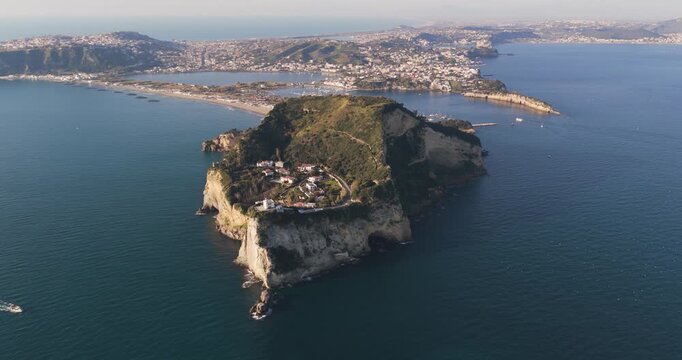 Aerial view of the dramatic cliffs of Capo Miseno meeting the azure sea, contrasting with the distant cityscape, creating a striking visual, Capo Miseno, Campania, Italy.
