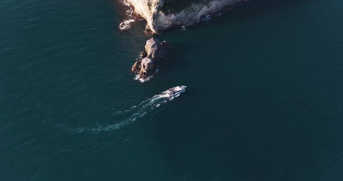 Aerial view of a boat sailing through the dark blue sea, contrasted by the white wake, near rocky cliffs, Capo Miseno, Campania, Italy.