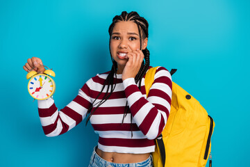 Young woman with striped top and yellow backpack holds alarm clock against blue background