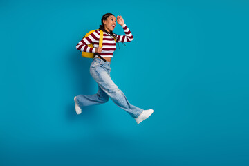 Young woman jumping with joy wearing striped top jeans and yellow backpack on blue background promoting active lifestyle and fashion
