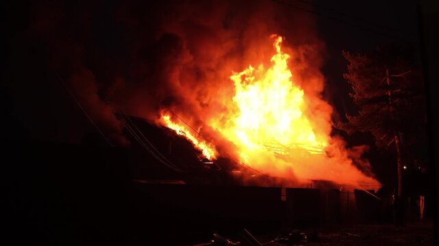 Large flames erupt from a house fire, illuminating the night sky and creating a dramatic scene in a quiet neighborhood.