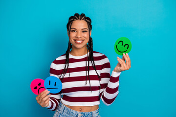 Young woman with braided hair smiles while holding colorful emoji signs in a bright blue studio...