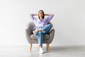 Happy peaceful young woman sitting in armchair with hands behind head, resting, having break from...