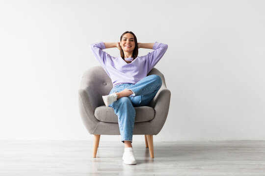 Happy peaceful young woman sitting in armchair with hands behind head, resting, having break from work against white studio wall, full length. Pretty lady chilling on weekend morning