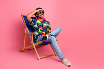 Fototapeta premium Young mixed race man relaxing on a blue deck chair with tropical lei sunglass and drink against pink background for a fun stylish leisure lifestyle image