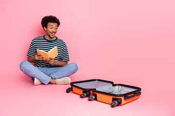 Young man reads a book while sitting on the pink studio floor beside an open orange suitcase creating a playful travel themed moment © deagreez