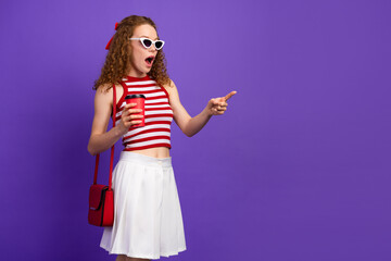 Young fashion model girl with red white striped top white skirt sunglasses holding a coffee cup points excitedly against a purple background