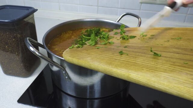Adding chopped fresh parsley from a wooden board into a boiling pot of traditional Ukrainian red borscht soup on an electric stove