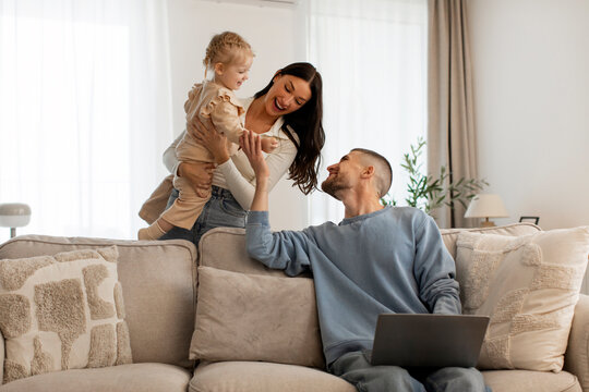 Mother carrying infant daughter father using laptop, working remotely from home and spending time with family