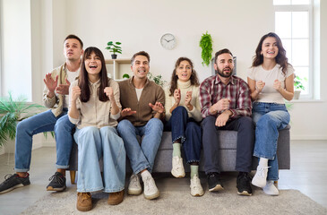 Friends cheering football on TV at home. Seven friends sit on a couch, clapping and pumping fists while watching a tense match in a bright living room. Joyful home sports viewing party.