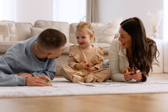 Loving parents chatting with their cute daughter while drawing together, lying on floor carpet in living room