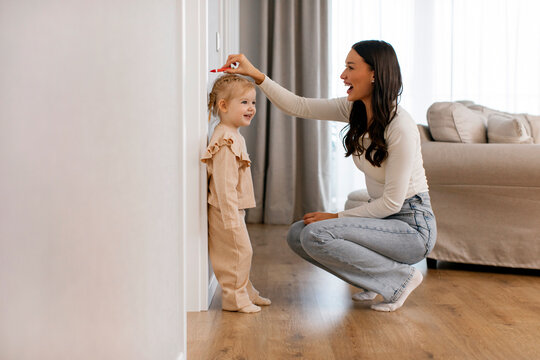 Kids Height Measurement. Loving mother measuring height of her little daughter near white wall in living room, side view